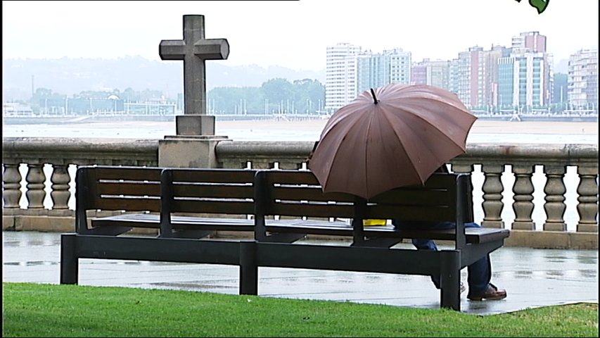 Una persona sentada en un banco con su paraguas frente a la Playa de San Lorenzo